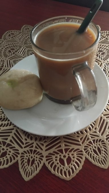 A glass mug of milky brown coffee with a spoon, served alongside a small pastry on a white plate over a decorative doily.
