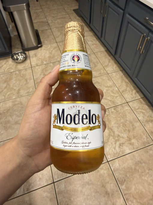 A hand holds up a bottle of **Modelo Especial** (Cerveza, since 1925) in a kitchen, the golden liquid and gold foil cap catching the light. The image pairs with the caption "time to speak my truth about artificial inu" — suggesting a relaxed, honest moment over a Mexican lager.