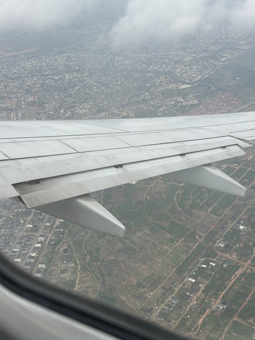 A view from an airplane window shows the plane's wing and flaps extended over a cloudy landscape of fields, roads, and scattered buildings below.