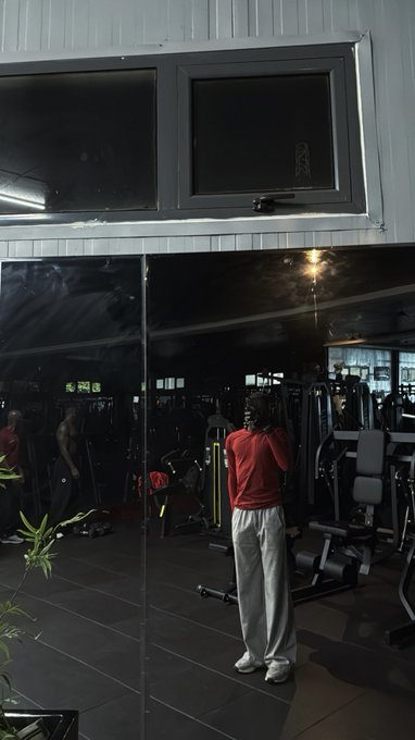 A man in a red shirt and gray sweatpants stands in a dimly lit gym with various workout equipment visible behind him, viewed through a glass window.