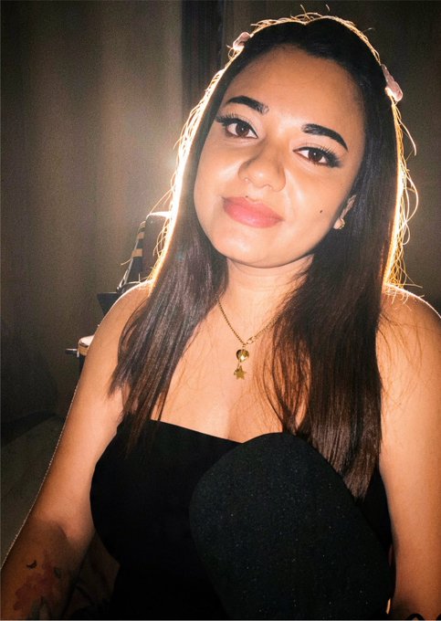 A portrait of a young woman with long dark hair wearing a black strapless top, posing with a slight smile in a dimly lit room.