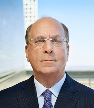 A professional headshot of a bald man with glasses wearing a dark pinstripe suit and blue patterned tie, photographed against a blurred modern building backdrop.