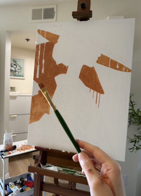 A hand holding a paintbrush in front of an easel displaying a canvas with brown paint strokes and pencil sketches, set in a bright art studio.