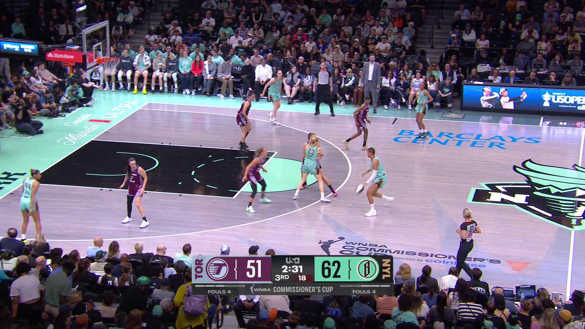 The image shows a basketball game taking place, with players actively engaged in the action on the court. The crowd of spectators can be seen watching the game from various angles around the court. There are at least 13 people visible in the scene, some standing closer to the court while others are further away. A sports ball is located near one of the players, indicating that they are currently involved in a play or have just finished one. The image also includes text elements such as "w nba.com/how-to-watch-g" and "TOR-NYL", which may be related to the basketball game being advertised on the website.