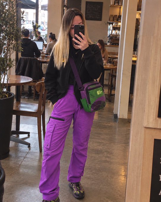Mirror selfie of a woman in a cafe wearing a black hoodie, bright purple cargo pants, and a purple and green North Face crossbody bag, taking the photo with her smartphone.