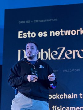 A man with a beard wearing a black hoodie stands on stage speaking at an event, with a screen behind him displaying "CABO 02 - INFRAESTRUCTURA" and "Esto es network... DoubleZero" along with other text about blockchain and validators.