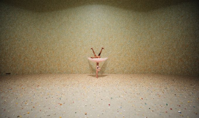 A small ballerina figurine in a pink tutu performing a handstand pose against a textured wall in a softly lit room with confetti-strewn floor.