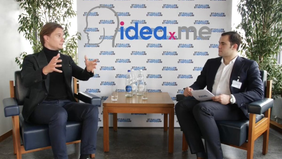The image shows a man sitting in a chair talking to another man who is also seated in a chair. They are both facing each other with their hands outstretched as if they're engaged in conversation or presenting something. The background features a white wall adorned with blue and gray logos, including one that reads "IDEA XME". There's also a table between the two men, which has several bottles on it.