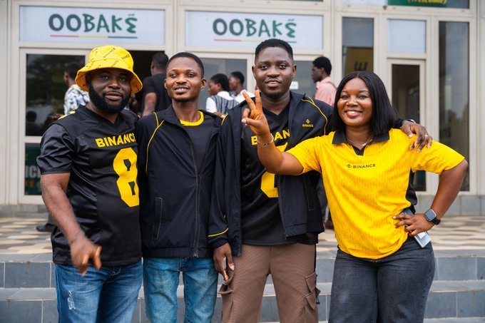 Four people pose in front of a building labeled 'OOBAKE', wearing black and yellow shirts with 'BINANCE' and '8' printed on them.