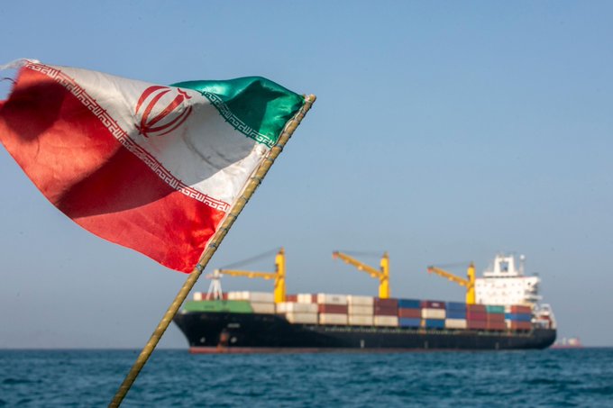 The image shows a large ship sailing across the ocean with Iran's flag flying from its mast. The ship appears to be a cargo vessel, as there are multiple containers visible on its deck. In the background, another ship can be seen further out in the water. The scene also includes a post that reads "BREAKING: Iran is reportedly turning the Strait of Hormuz into a money-printing machine." This suggests that Iran's maritime activities have become a significant source of revenue for them through the use of cryptocurrency and digital payments, as indicated by the post.