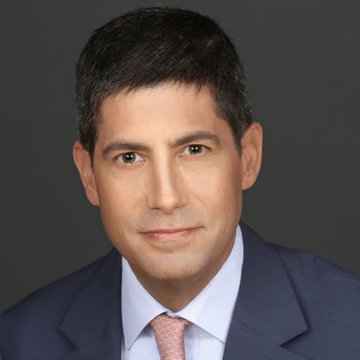 A professional headshot of a man with dark hair wearing a navy blue suit, light blue shirt, and pink patterned tie against a gray background.