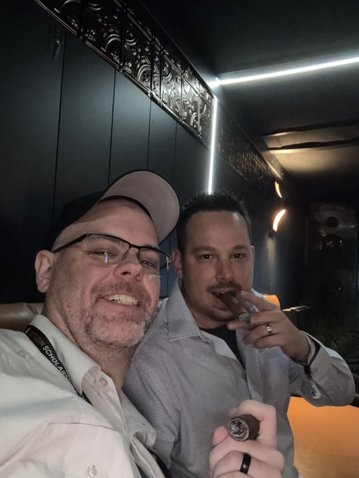 Two men wearing caps and collared shirts are smoking cigars in a dimly lit room.