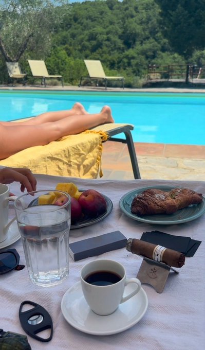 Poolside breakfast scene with a cup of coffee, water, fruit, croissants, and a cigar on a table; a person reclines on a lounge chair beside a swimming pool.