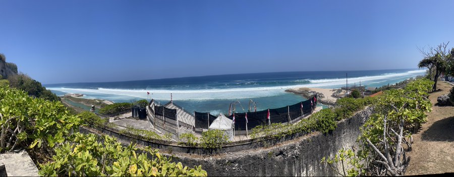 A scenic coastal view shows a beach, ocean waves, and a fenced event area under a clear blue sky.
