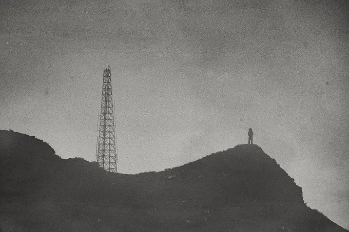 A grainy black-and-white photograph showing a silhouetted figure standing on a hill next to a tall transmission tower.
