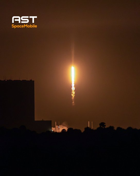 A nighttime rocket launch ascends with a bright orange flame trailing from its engines, silhouetted against the dark sky above a distant launch facility. The image features the "AST SpaceMobile" logo in the top left corner, reinforcing the company's branding alongside the $ASTS ticker mentioned in the post.