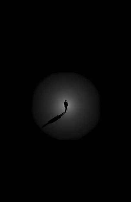 A silhouette of a solitary person standing in a circular pool of light against a dark background, casting a long shadow.