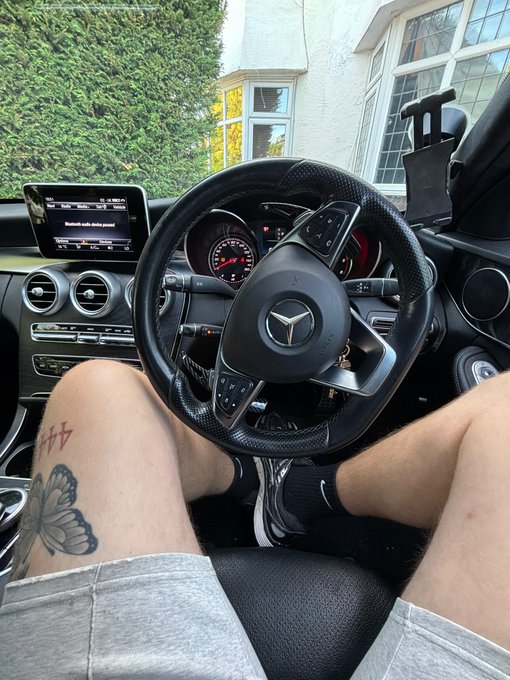 A first-person perspective from the driver's seat of a Mercedes-Benz vehicle, showing a person's legs with butterfly and cross tattoos, the steering wheel, and the dashboard infotainment system.