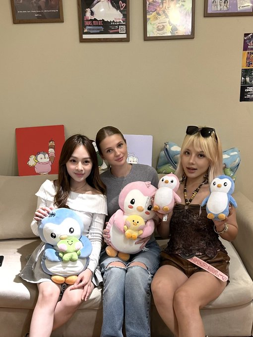 Three young women sit on a beige couch holding plush penguin and frog toys, with framed posters on the wall behind them including one advertising a 'TOURS' event.