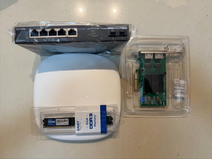 Networking equipment laid out on a counter: a Tenda SG2007X switch, a white device (possibly a base station), an Expert DDR3 RAM module in packaging, and a green circuit board adapter in a clamshell case.