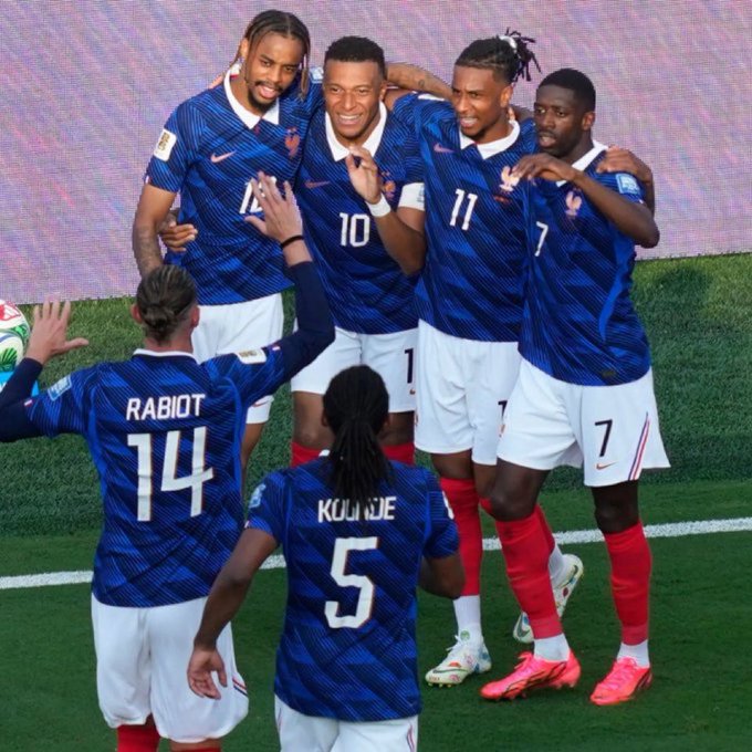 French national soccer players, including Mbappé (number 10), Rabiot (number 14), and Koundé (number 5), celebrate together on the field wearing their blue jerseys.