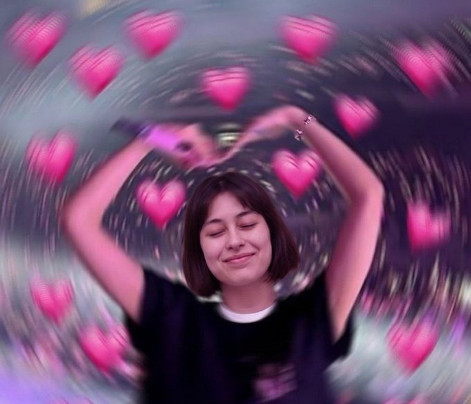 A young woman with short brown hair smiles with her eyes closed and arms raised, surrounded by an animated swirl of glowing pink hearts.