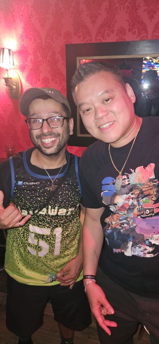 Two men posing together and smiling at the camera, one wearing glasses, a cap, and a green basketball jersey, the other in a black graphic t-shirt, standing in front of a red patterned wall.
