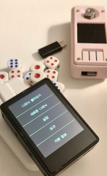 A handheld device displays a menu in Korean with options including "니모닉 불러오기" (Load mnemonic), "새로운 니모닉" (New mnemonic), "설정" (Settings), "도구" (Tools), and "기본 정보" (About), accompanied by a pile of six-sided dice and a pink SeedSigner-style hardware device with an empty entropy entry slot—visually reinforcing the comparison between Krux and SeedSigner dice-roll mnemonic generation mentioned in the post.