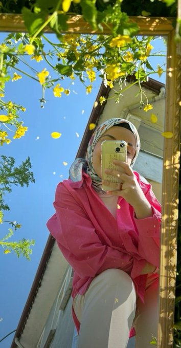 A mirror selfie of a person wearing a pink blouse and patterned headscarf, taken outdoors against a bright blue sky framed by green foliage.