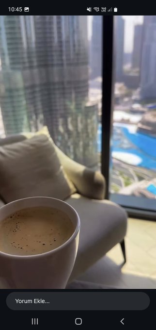 A first-person photo taken from a high-rise balcony shows a cup of coffee in the foreground with a view of skyscrapers and a pool area below, taken at 10:45.