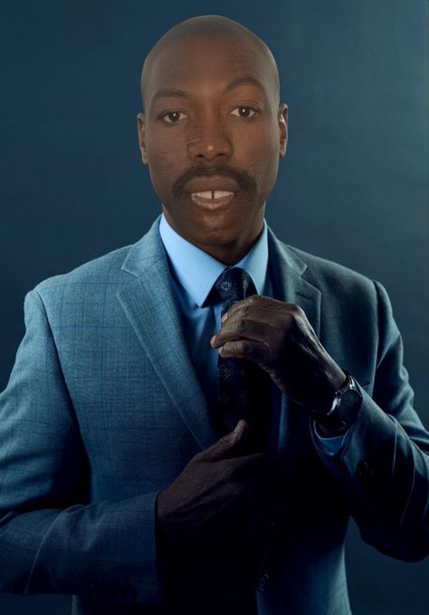 A man in a blue suit and tie adjusts his cuff against a dark blue studio background.