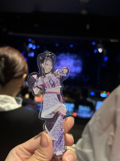 A hand holding up an acrylic standee of an anime girl character wearing a purple and white outfit at what appears to be a concert venue.