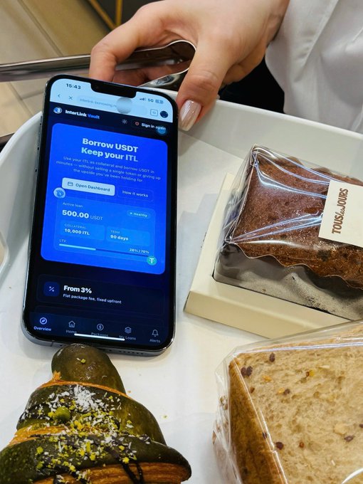 A smartphone displays the InterLink Vault interface showing an offer to "Borrow USDT Keep your ITL," with options for "Open Dashboard" or "How it works," alongside an "Active loan" of 500.00 USDT against 16,000 ITL collateral—demonstrating the collateral-based borrowing dApp in action right next to some pastries.