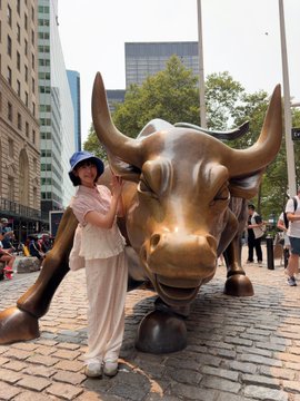 A woman wearing a blue hat and beige outfit poses next to the iconic Charging Bull statue in New York City's Financial District.