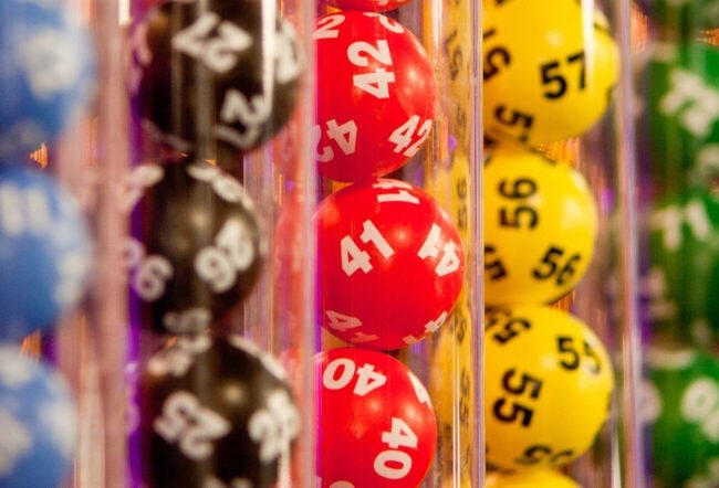 Close-up of lottery balls in red, yellow, green, and black tubes with numbers like 40, 41, 42, 55, 56, 57 visible.