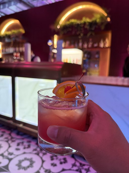 A hand holding a pink cocktail garnished with an orange slice and a maraschino cherry in a rocks glass, set against the backdrop of a dimly lit bar with glowing arches and shelves of liquor bottles.