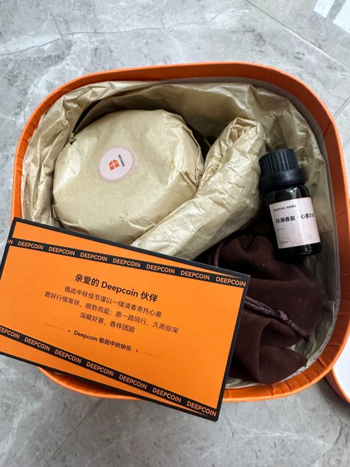 An orange Deepcoin promotional gift box containing a round wrapped item, a small bottle of essential oil, a scarf, and a card with Chinese text about Deepcoin's values.