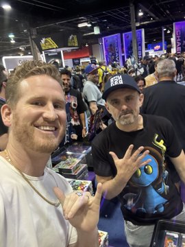 Two men pose for a photo at a bustling convention, with the man on the right wearing a black cap reading "VEEFRIENDS" and a matching t-shirt featuring the brand's colorful character designs. The image captures the meet-and-greet moment at the VeeFriends booth referenced in the caption, where fans gathered around to get autographs and interact with Gary Vaynerchuk.