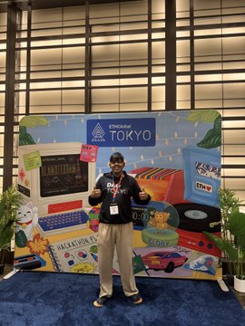 A man posing with two thumbs up in front of a 'Beautiful TO'KYO' promotional photo backdrop featuring pop culture and retro Japanese imagery, taken at what appears to be a conference or event.
