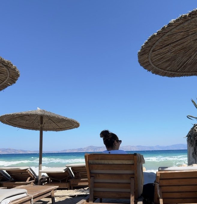 A woman relaxes on a beach lounger under thatched umbrellas, looking out at the turquoise ocean and distant mountains under a clear blue sky.