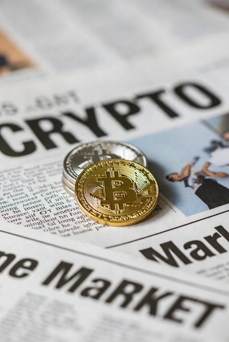 A close-up photograph of a gold Bitcoin coin resting on top of a silver coin, placed on a newspaper page featuring the large words "CRYPTO" and "the Market."