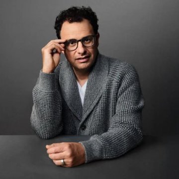 Dario Amodei, CEO of Anthropic, wearing glasses and a gray cardigan, rests his hand on his face in a thoughtful pose. The image accompanies news of his announcement about Claude AI's breakthrough in identifying a novel enzyme system in bacteriophage DNA.