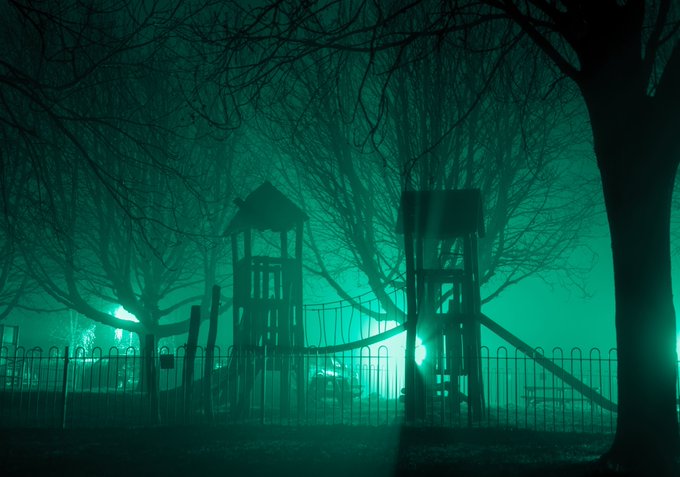 An eerie, green-tinted nighttime photograph of a playground with two tower structures, a slide, and a metal fence, surrounded by silhouetted bare trees and fog.