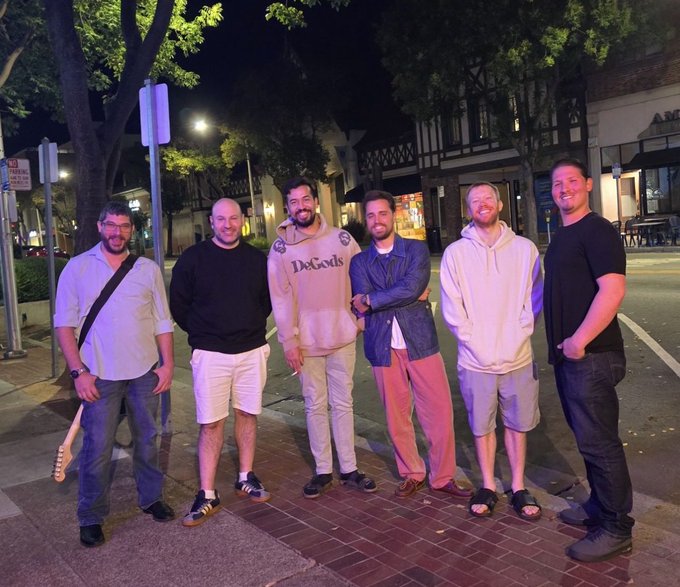 Six men stand smiling together for a group photo on a sidewalk at night in front of a Tudor-style building, with one wearing a hoodie that reads 'DeGods'.