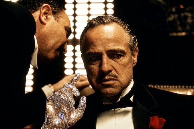 A cinematic portrait of an older man in a black tuxedo with a red pocket square and a bow tie, shown with another man whispering in his ear, reminiscent of The Godfather film.