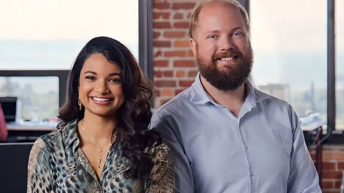 A professional portrait of a smiling man and woman standing side-by-side in an office setting with an exposed brick wall and large windows in the background.
