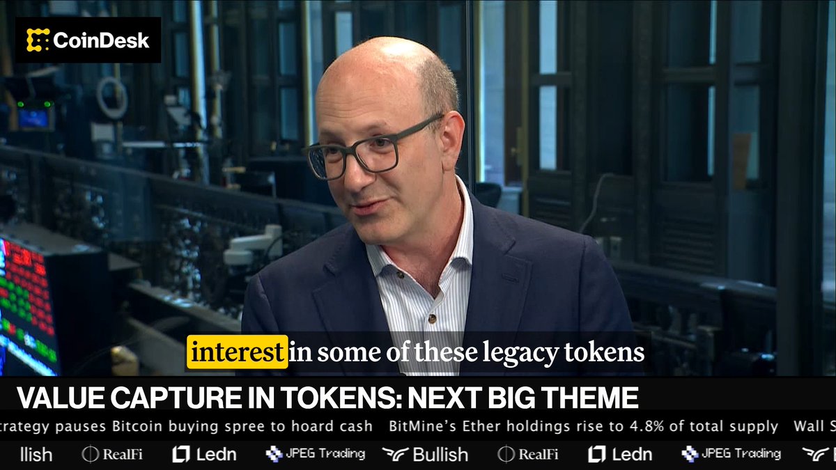 A bald man in glasses and a dark suit speaks in a control room-style setting with monitors displaying data, while the CoinDesk ticker at the bottom reads "VALUE CAPTURE IN TOKENS: NEXT BIG THEME" with a highlighted caption noting "interest in some of these legacy tokens," visually illustrating the discussion about a resurgence in established crypto projects.