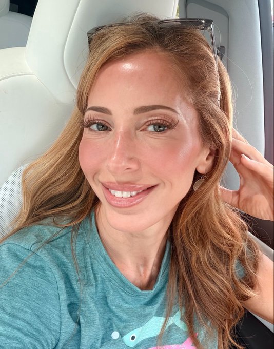 A close-up selfie of a smiling woman with long strawberry-blonde hair wearing sunglasses on her head, sitting in what appears to be an airplane seat.