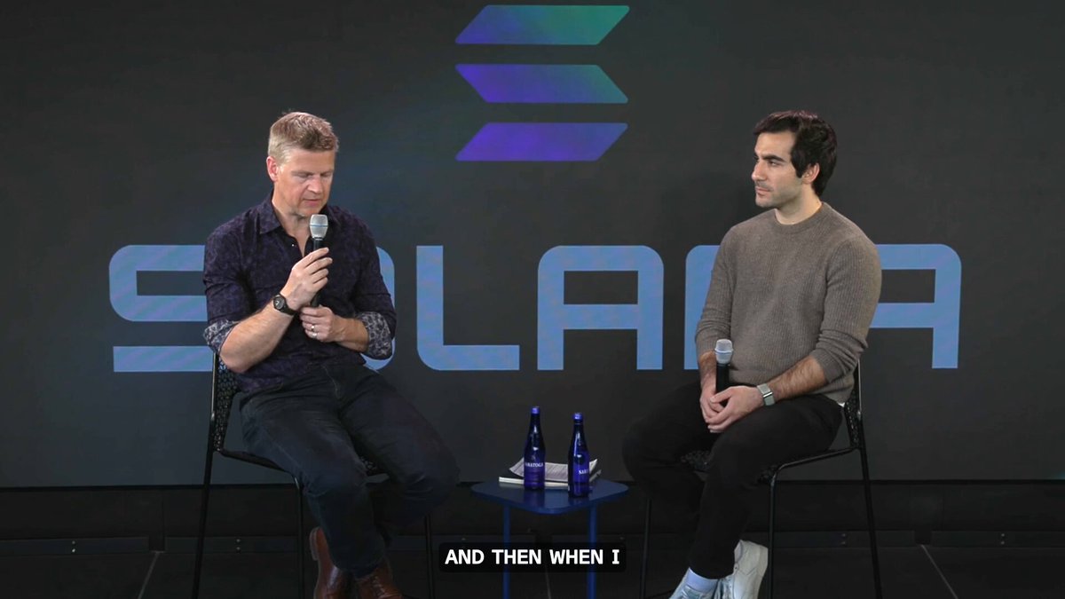 The image features two men sitting next to each other at a table with microphones, appearing to be engaged in conversation or participating in an interview. The background of the image includes a large blue sign that says "Solana" written in white letters. There are also several bottles placed on the table between the two men.