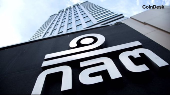 A low-angle shot shows a tall office building towering behind a large black sign displaying the white "i am" logo. The image, branded with the CoinDesk logo, accompanies news that Thailand's SEC has approved domestic Bitcoin ($BTC) and Ethereum ($ETH) ETFs, though no specific figures or tickers appear in the scene itself.